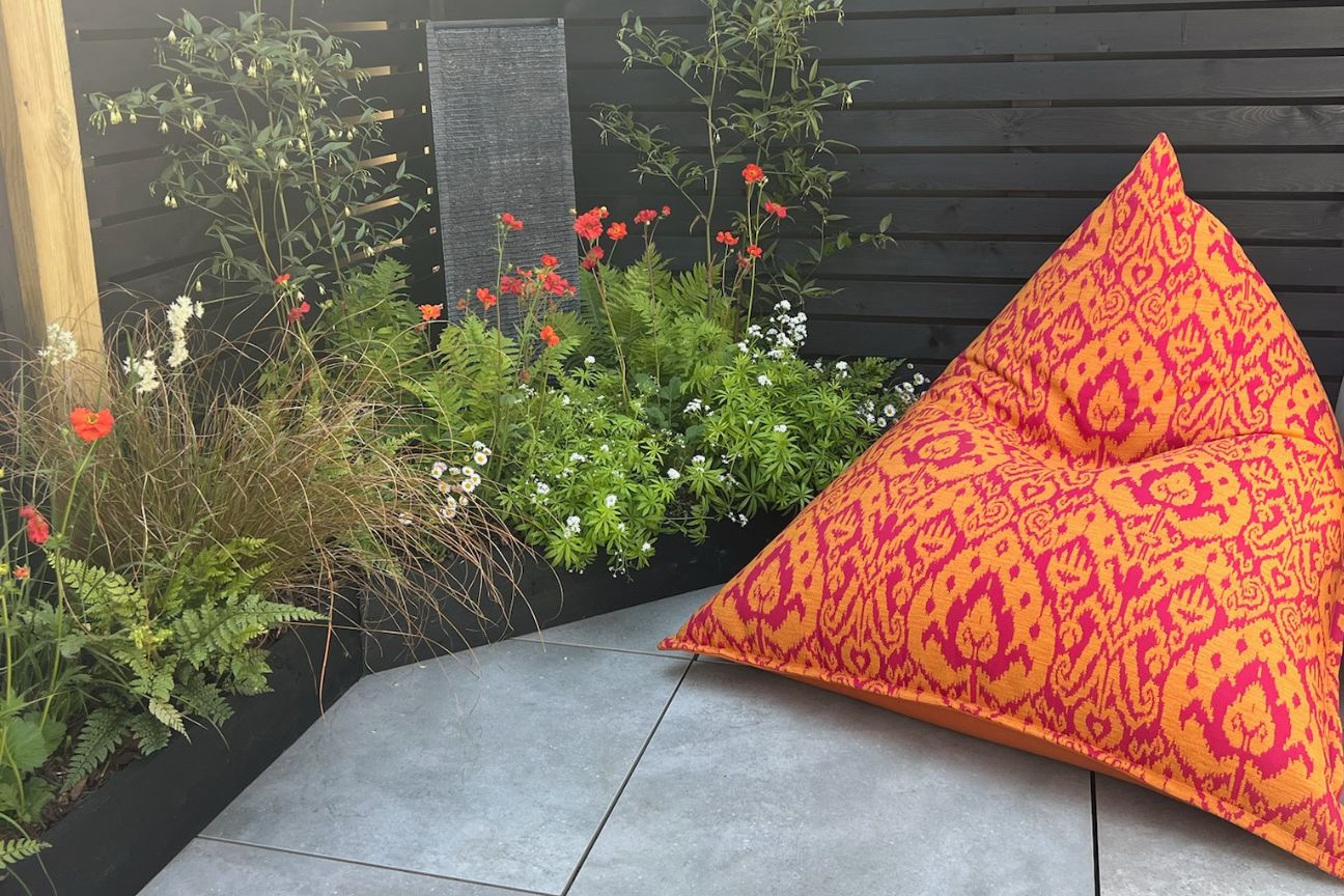 Brightly coloured garden bean bag chair in orange patterned fabric contrasing with a black painted fence, green foliage, and bright red geums.