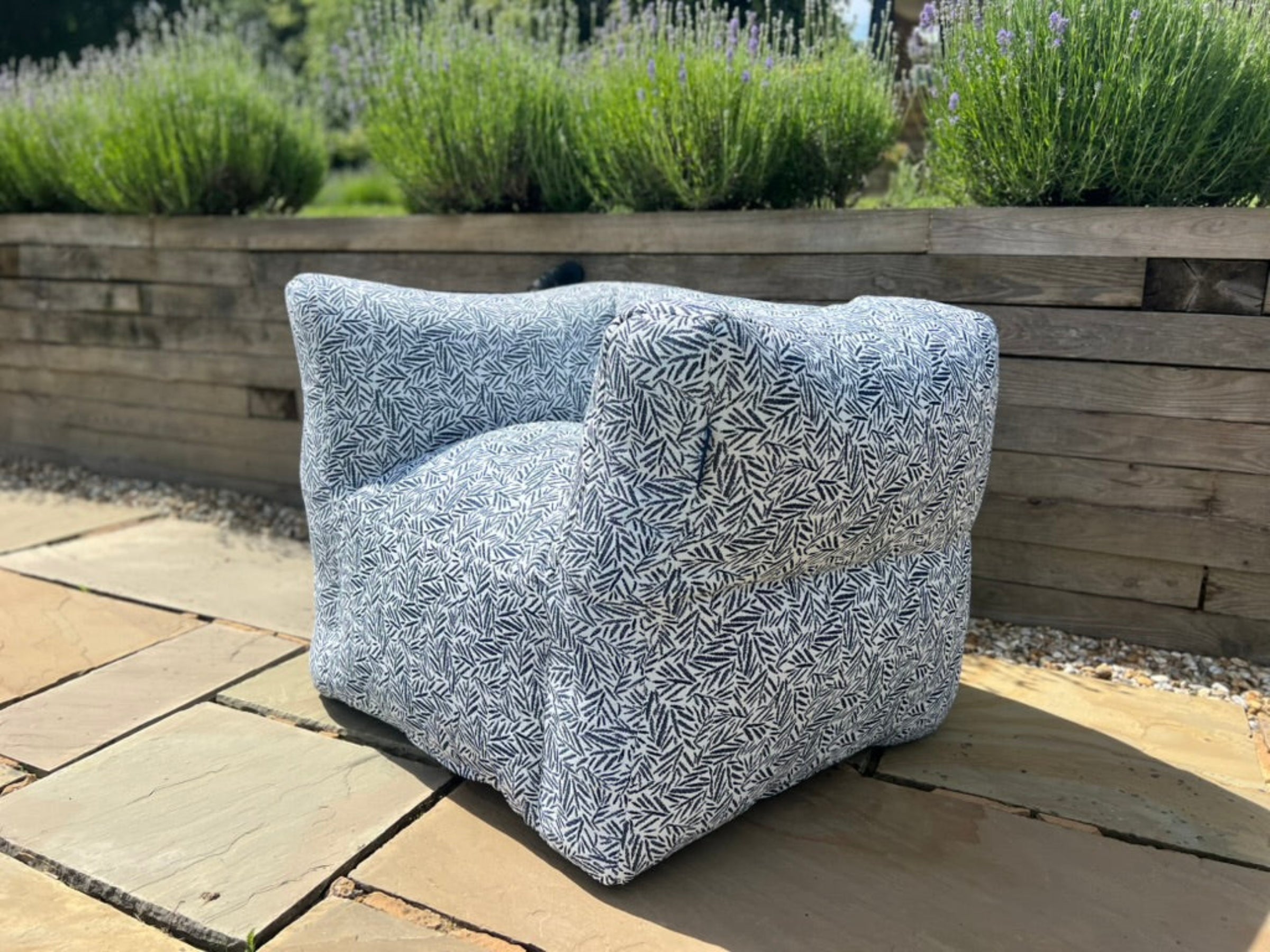 A modern low-profile outdoor bean bag armchair with a dense blue and white abstract leaf-like patterned fabric, set outdoors on a stone patio with a wooden planter wall and greenery in the background.