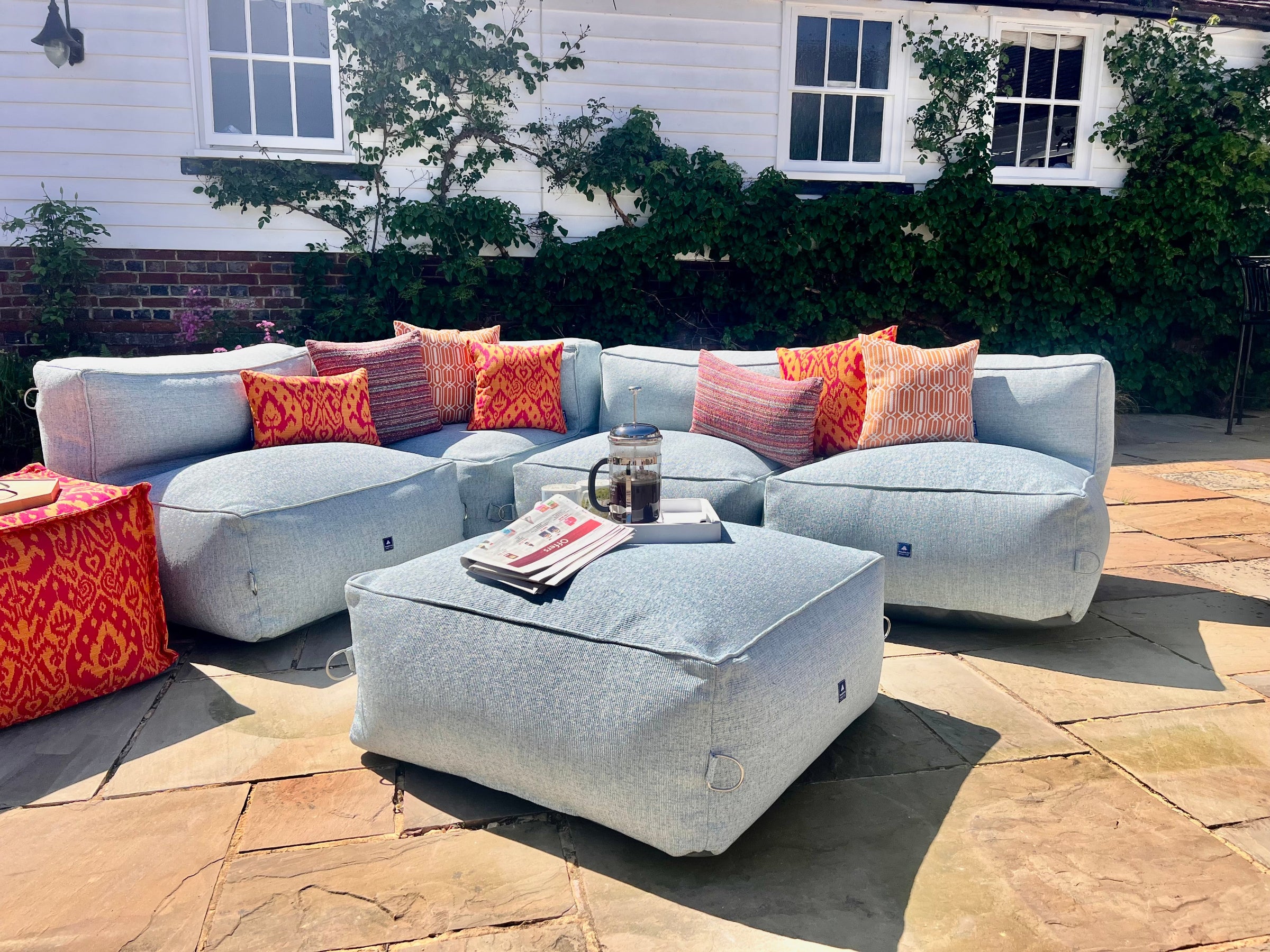 Outdoor bean bag modular sofa set in light blue textured material called sea spray on a stone patio in front of a white house. featuring red and orange medina, calypso and ripple patterned outdoor cushions and a matching ottoman coffee table and pouffe