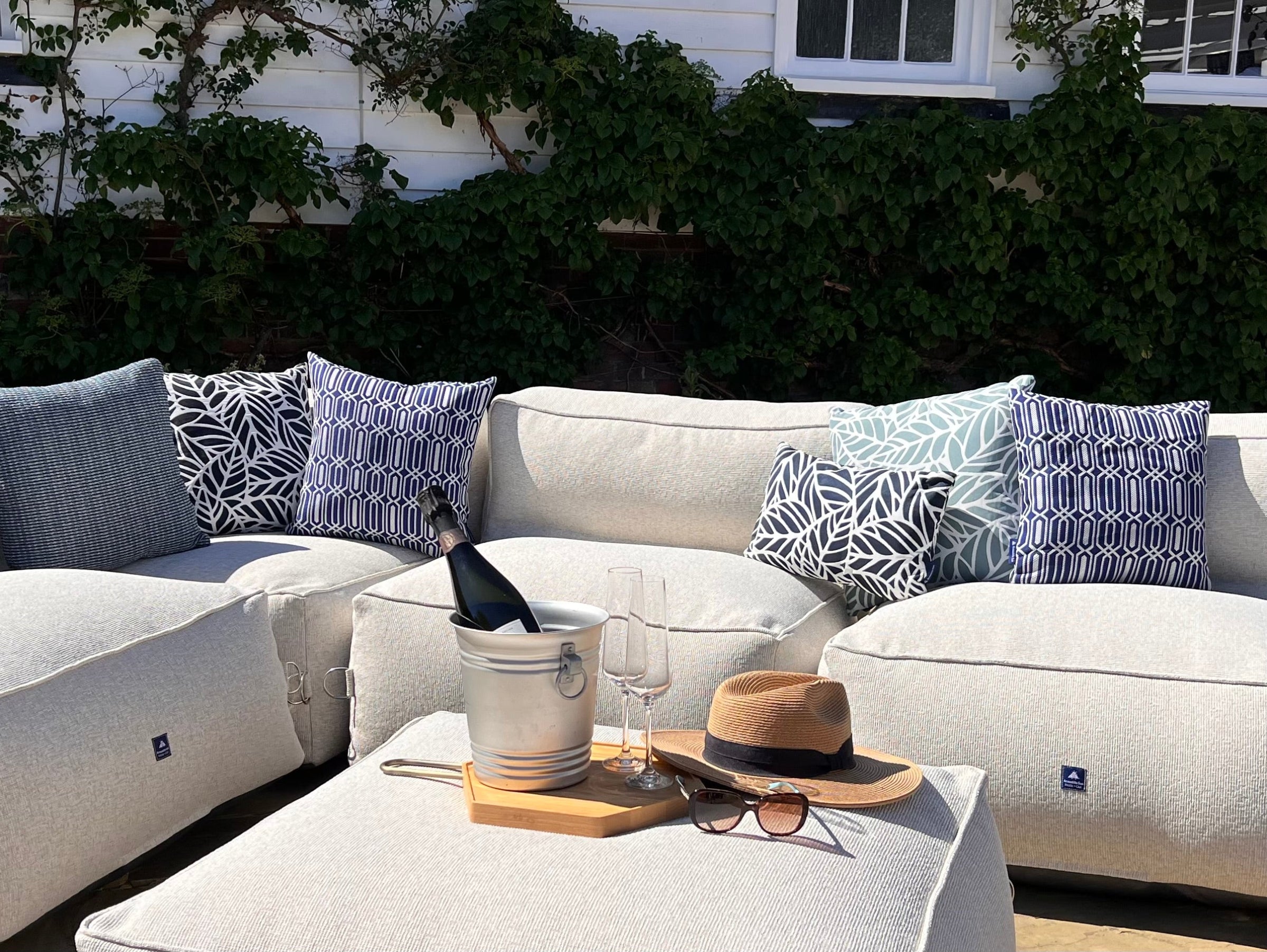 Outdoor beanbag sofa set in natural shell, with calypso, palm and woven patterned blue cushions, a matching ottoman coffee table, a silver ice bucket with a wine bottle, two wine glasses, a tan hat, and sunglasses, set against a background of green ivy and a white house.