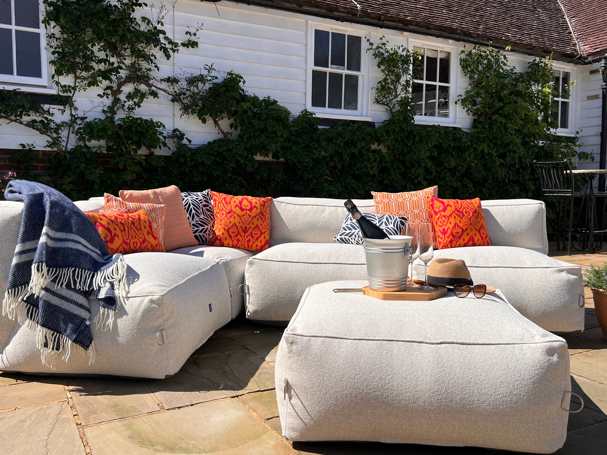 Outdoor bean bag modular sofa set in natural shell textured material with orange medina, calypso and pinstripe outdoor cushions and palm navy cushions. A blue fringed throw blanket, and a matching ottoman on a stone patio in front of a white house with green ivy.
