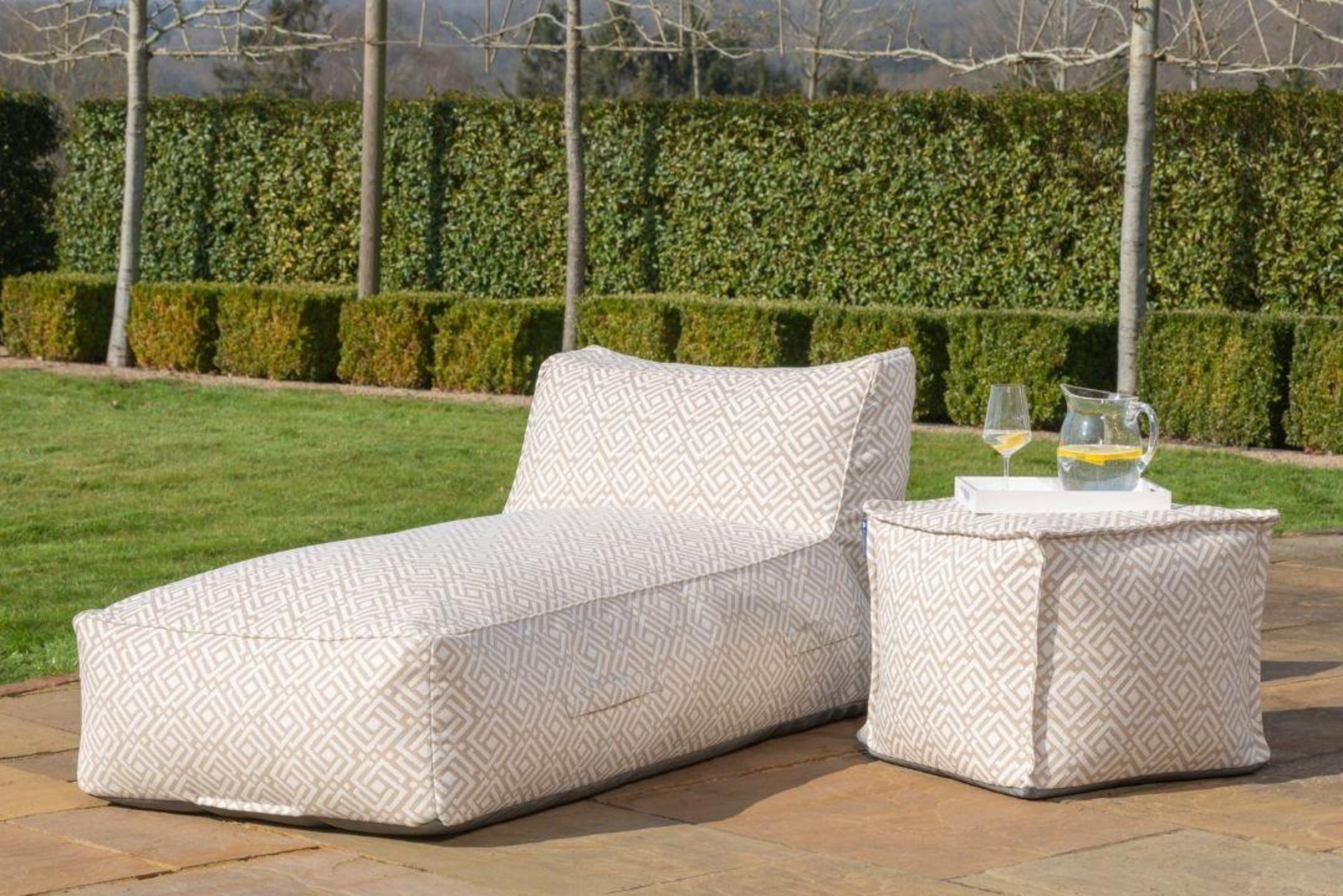 Outdoor bean bag sun lounger and matching side table in a garden setting, both upholstered in a light beige geometric fabric called sigma. The lounge is shown on a stone patio with a glass of wine and a pitcher on the side table, with a green hedge and lawn in the background.