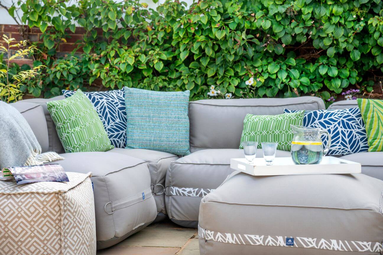 A cosy garden sofa on a private patio surrounded by green foliage and covered in outdoor cushions in blues and greens.