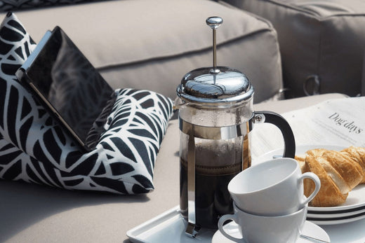 A bean bag iPad holder propping a tablet up securely outdoors next to a cafetiere and inviting plate of pastries.