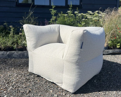 A modern, low-profile outdoor armchair in a textured light beige fabric, positioned on a gravel surface with a garden and dark blue house in the background.