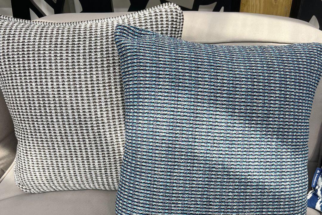 Two luxury outdoor cushions in a woven waffle-textured fabric. One is light charcoal grey with a darker grey texture; the other has a turquoise and grey-blue background with a dark blue-grey texture.