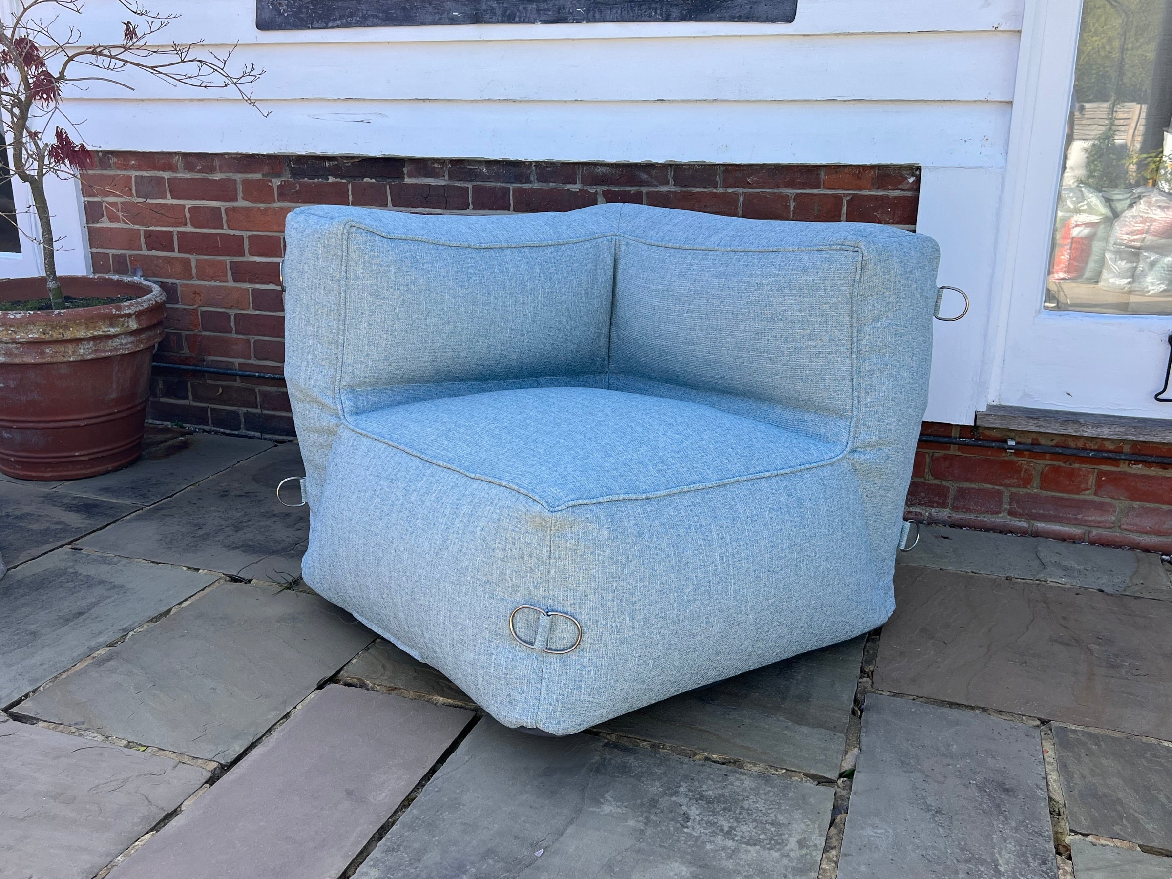 An outdoor bean bag corner chair for a sofa set in a light blue textured material called sea spray armchair on a stone patio with a brick wall and door in the background