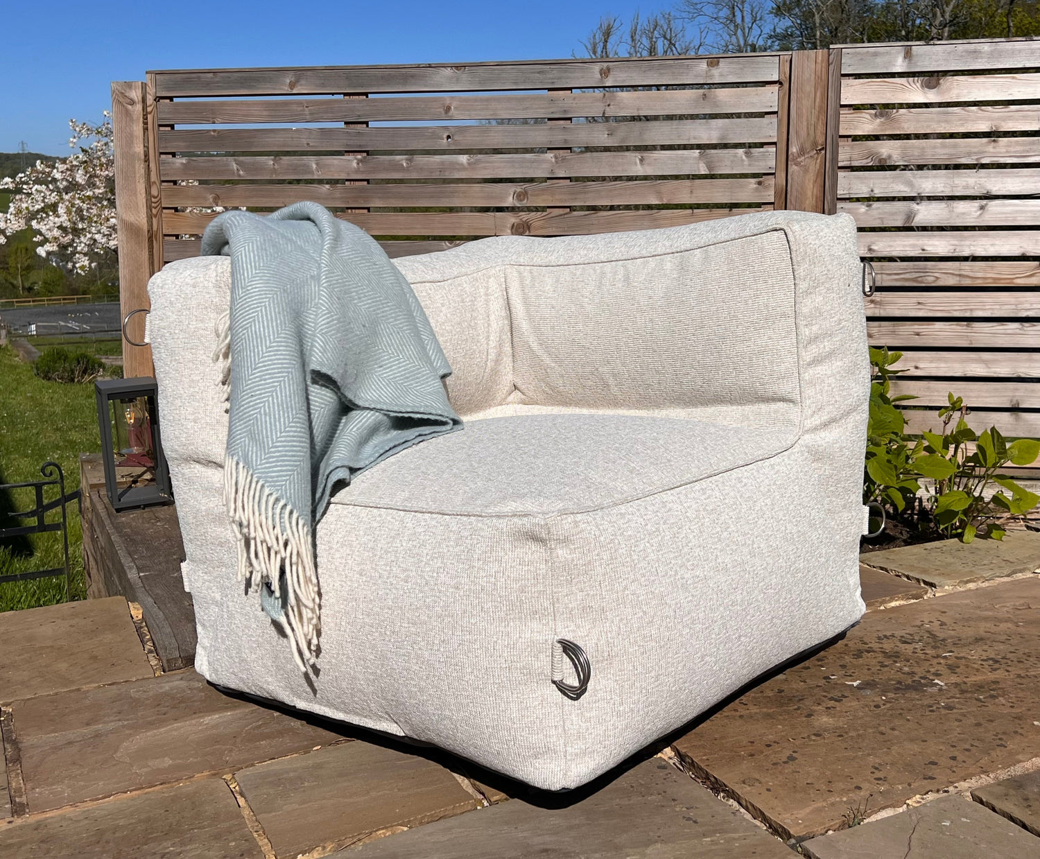 A natural shell coloured outdoor bean bag corner module for a sofa with a light blue throw blanket draped over it, set on a stone patio in front of a wooden fence.