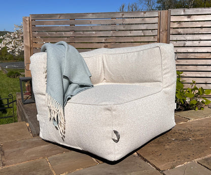 A natural shell coloured outdoor bean bag corner module for a sofa with a light blue throw blanket draped over it, set on a stone patio in front of a wooden fence.