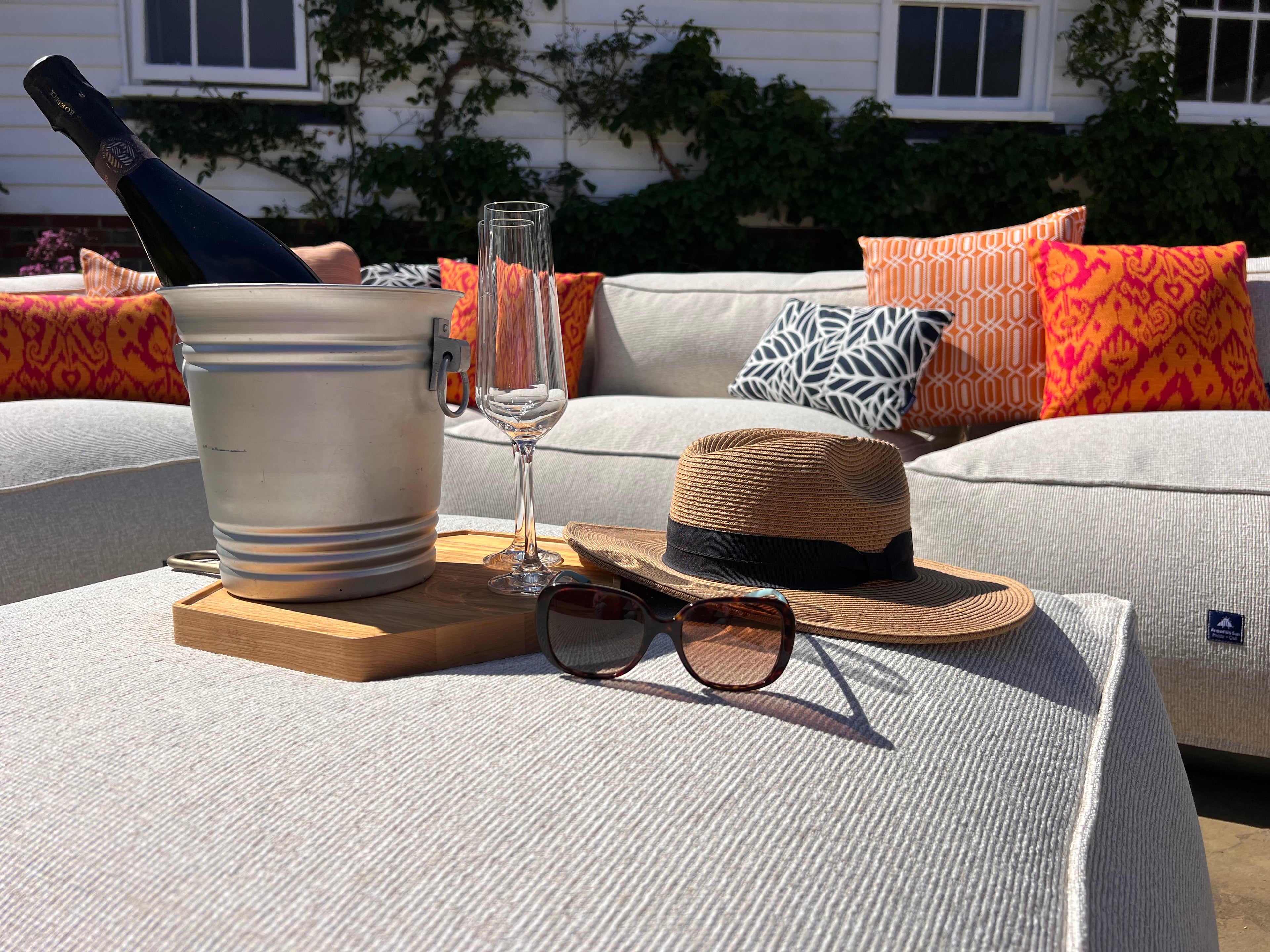 Outdoor bean bag sofa set in natural shell textured material with orange medina and calypso cushions pictured with a hat, sunglasses, and wine glass on a table with a house in the background.