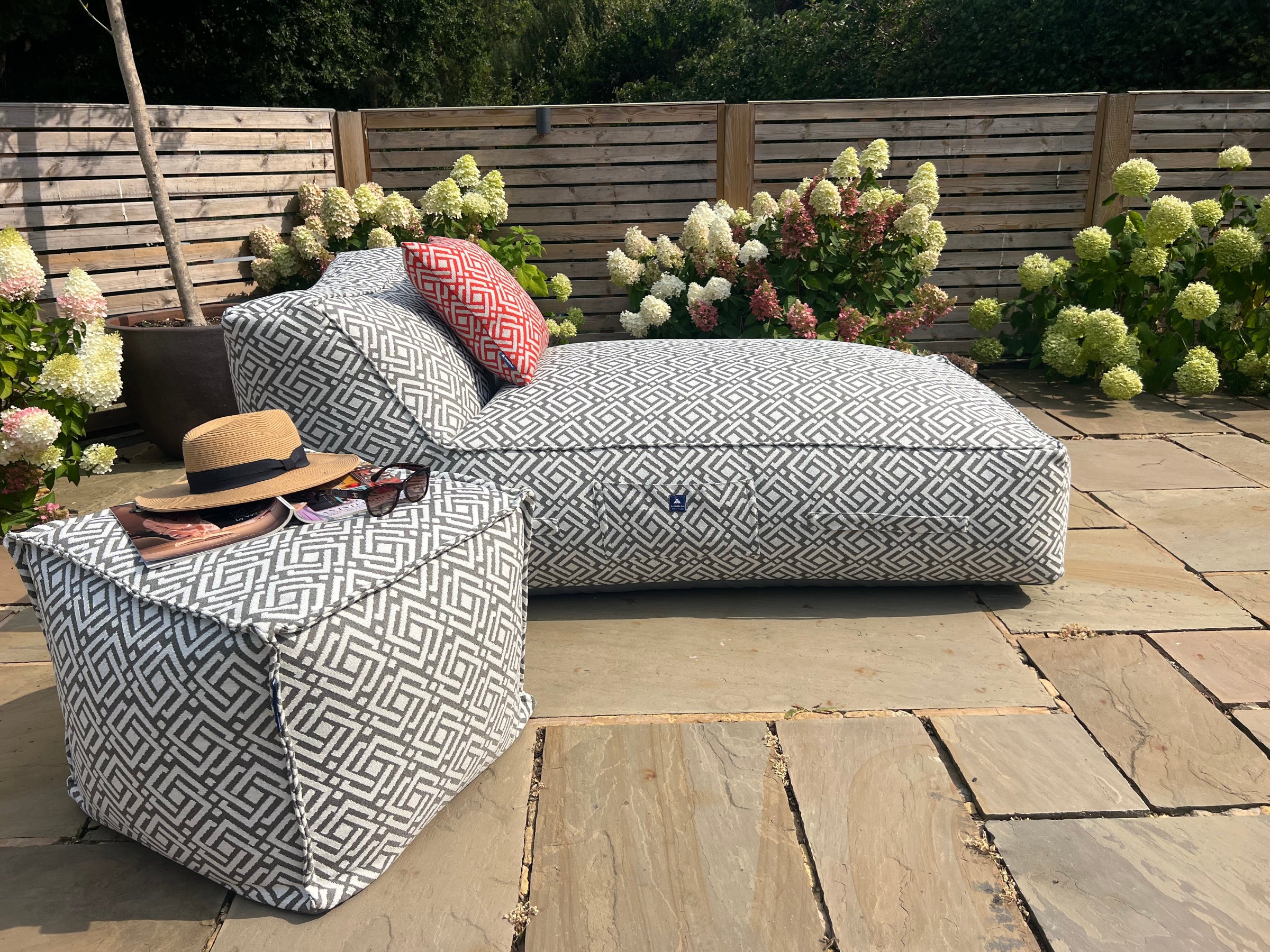 Outdoor bean bag sun lounger in geometric grey sigma pattern wih matching pouffe and cushions in paprika on a stone patio, surrounded by flowers and plants.