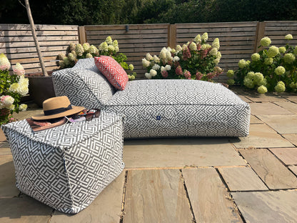 Outdoor bean bag sun lounger in geometric grey sigma pattern wih matching pouffe and cushions in paprika on a stone patio, surrounded by flowers and plants.