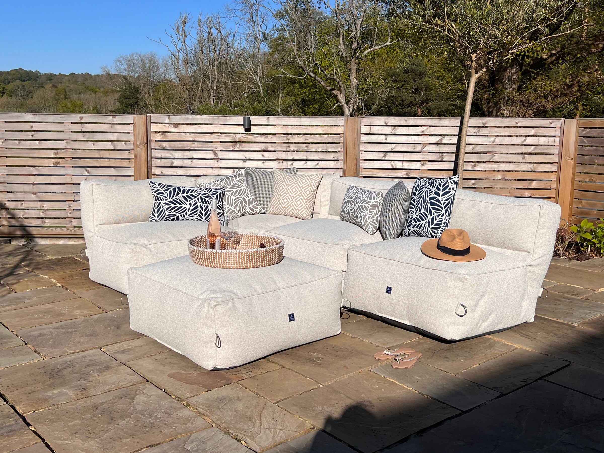 Outdoor bean bag modular sofa set in light natural textured fabric called shell on a stone patio, with matching ottoman, a woven tray table, and a straw hat in a backyard setting. Matched with palm navy, natural sigma, palm pumice, and waffle outdoor cushions.