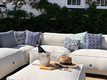 Outdoor beanbag sofa set in natural shell, with calypso, palm and woven patterned blue cushions, a matching ottoman coffee table, a silver ice bucket with a wine bottle, two wine glasses, a tan hat, and sunglasses, set against a background of green ivy and a white house.