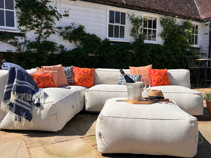 Outdoor bean bag modular sofa set in natural shell textured material with orange medina, calypso and pinstripe outdoor cushions and palm navy cushions. A blue fringed throw blanket, and a matching ottoman on a stone patio in front of a white house with green ivy.