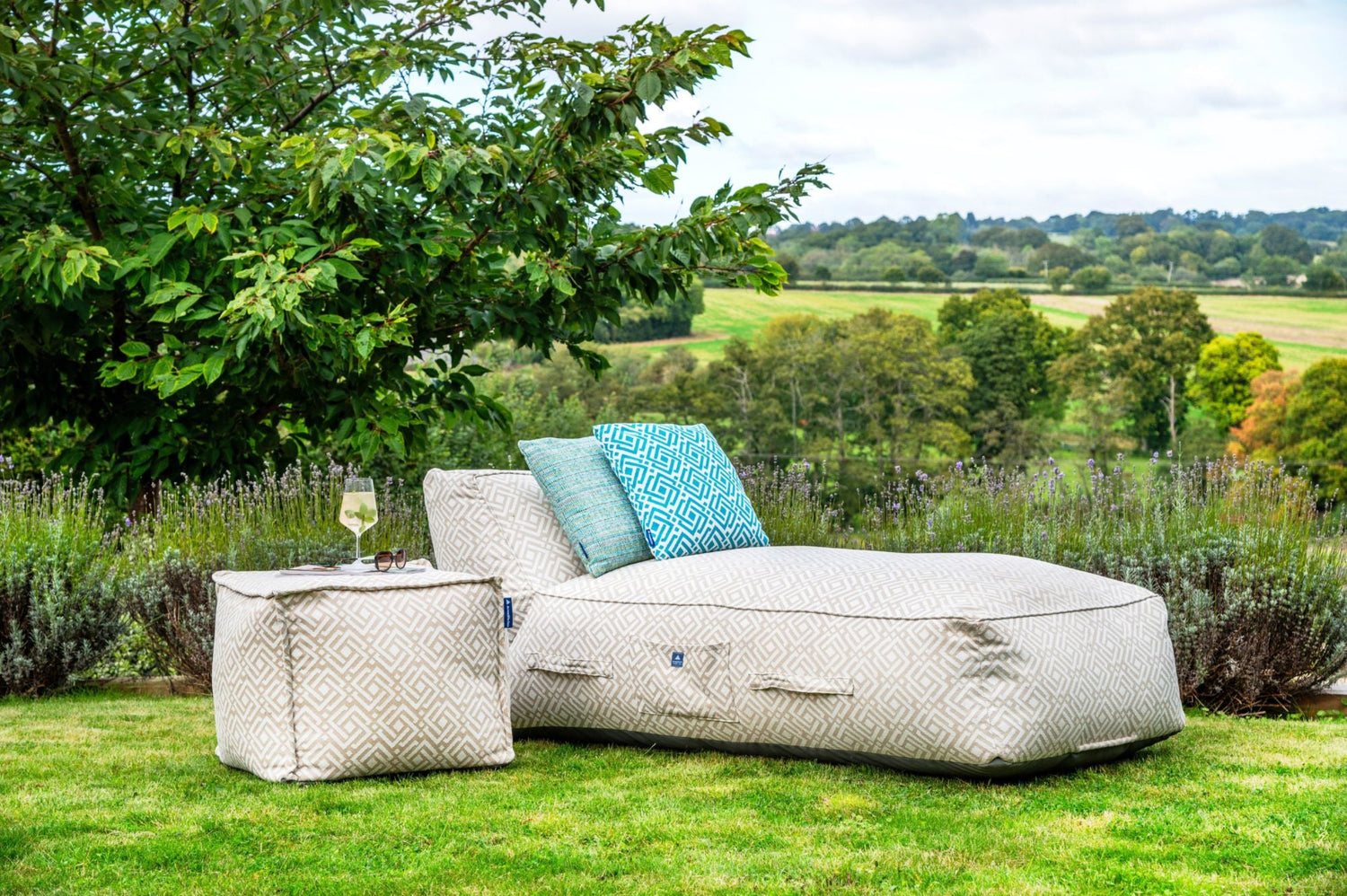 Outdoor bean bag sun lounger in geometric natural sigma patter with matching pouffe and outdoor cushions in turquoise on a grassy area with a scenic background