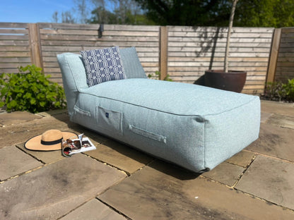 A light blue textured material outdoor bean bag lounge chair on a stone patio with a wooden fence and plants in the background. A Navy Calypso outdoor cushion,  a straw hat, and sunglasses are placed near the chair.