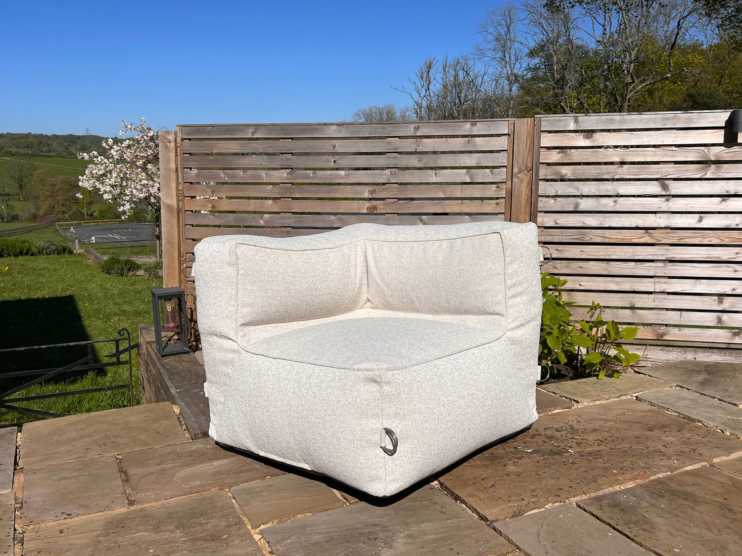 Natural shell coloured outdoor bean bag sofa corner module on a stone patio with wooden fence and trees in the background