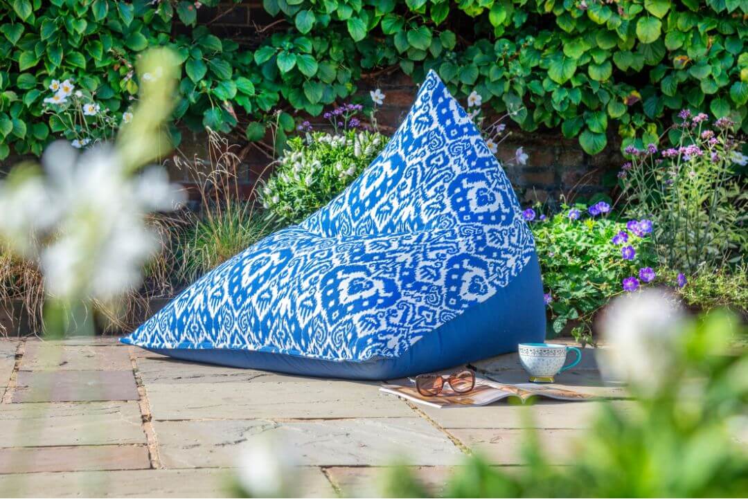 A blue and white patterned bean bag chair set invitingly on the patio amongst foliage and purple flowers.