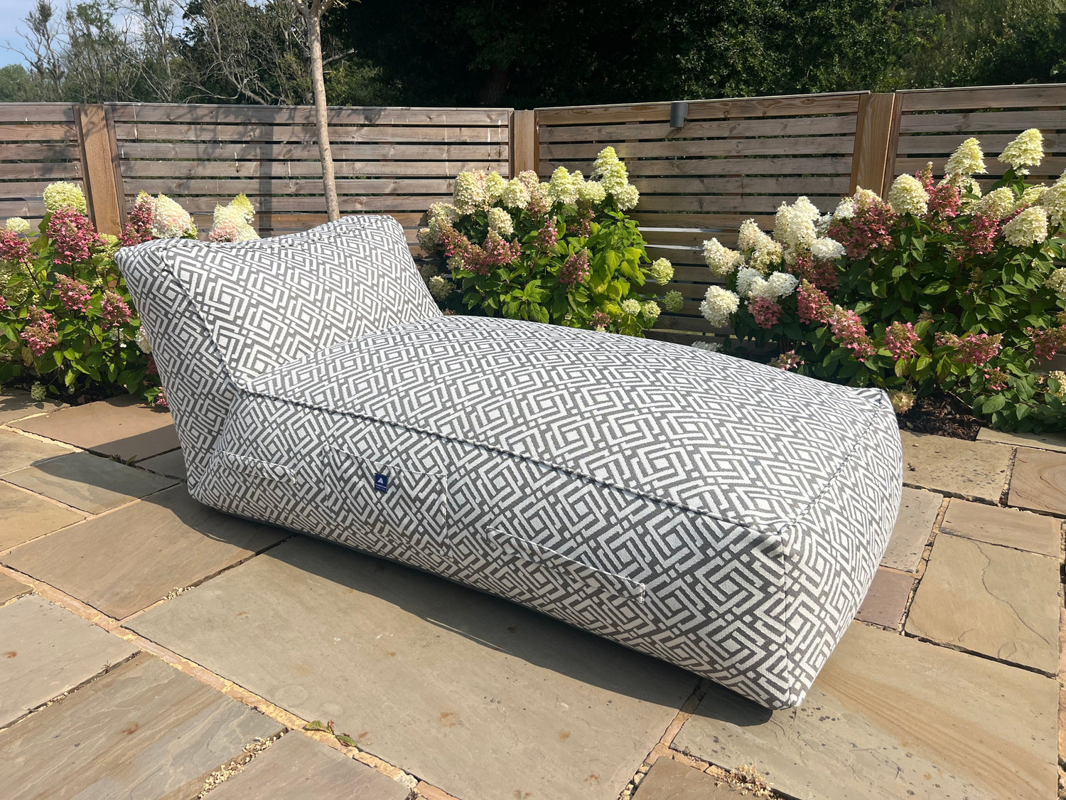 A grey geometric patterned bean bag sun lounger called sigma on a patio with flowers and wooden fence in the background