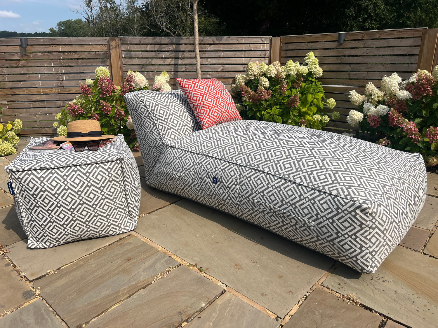 Outdoor bean bag sun lounger with geometric grey sigma pattern on a stone patio with flowers and a wooden fence in the background. accompanied with matching pouffe and cushion in paprika.