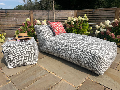 Outdoor bean bag sun lounger with geometric grey sigma pattern on a stone patio with flowers and a wooden fence in the background. accompanied with matching pouffe and cushion in paprika.