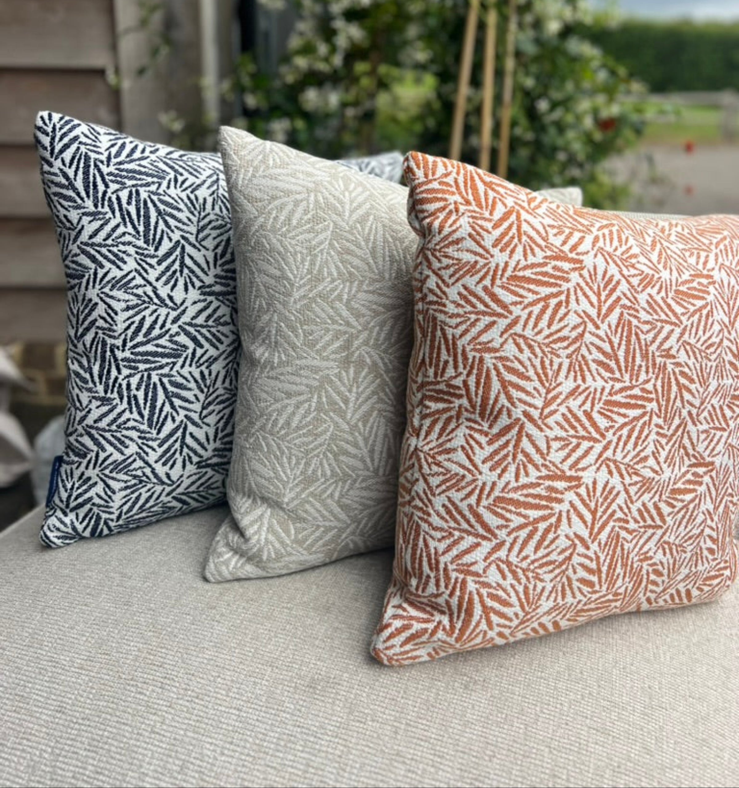 Three decorative outdoor cushions with botanical leaf patterns arranged on our shell outdoor sofa. The left pillow is navy blue and white, the middle pillow is natural and white, and the right pillow is terracotta orange and white.