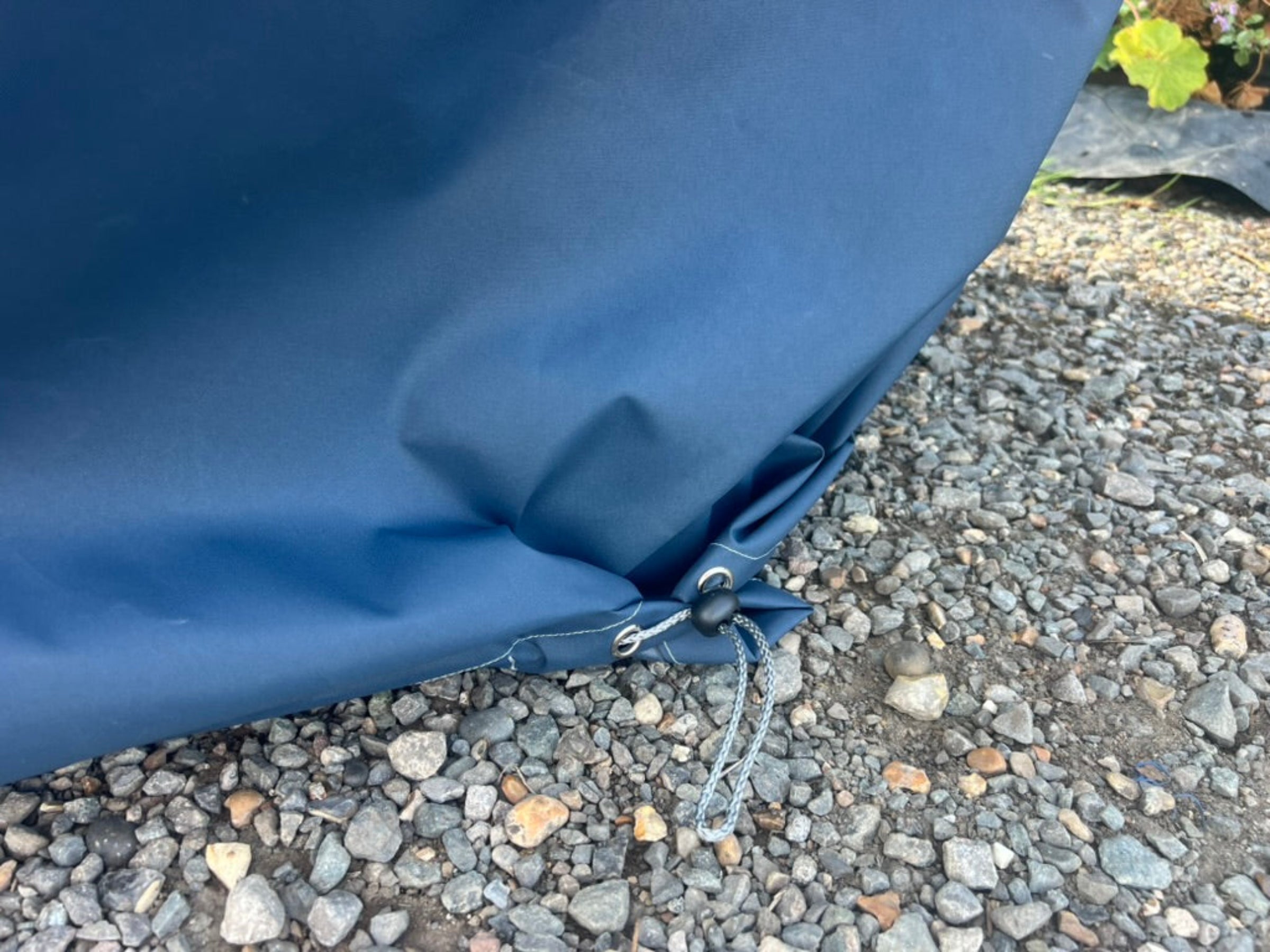 Close-up of a waterproof cover for modular corner chair or armchair for winter use. Secured with metal grommets and a drawstring cord, resting on gravel outdoors.