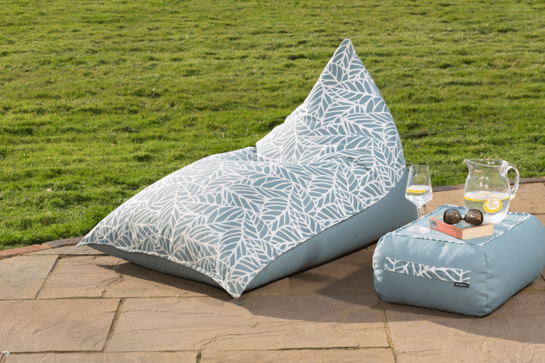 Outdoor bean bag lounger in blue palm pattern sits invitingly on a gardenpatio next to a table with a book and refreshing glass of water 
