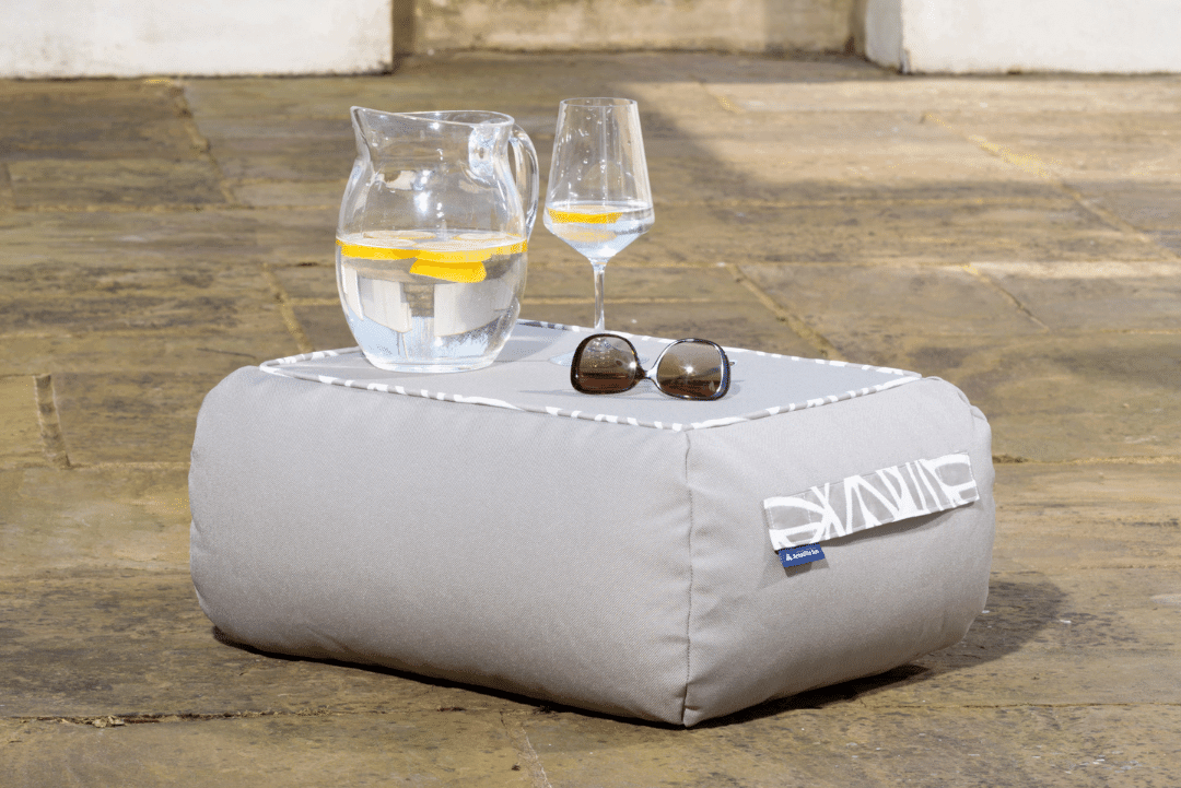 Grey bean bag coffee table with palm patterned detail around the edge of the top surface. Small and rectangular in shape with a flat base. Jug of water and a glass are placed on the top next to some sunglasses