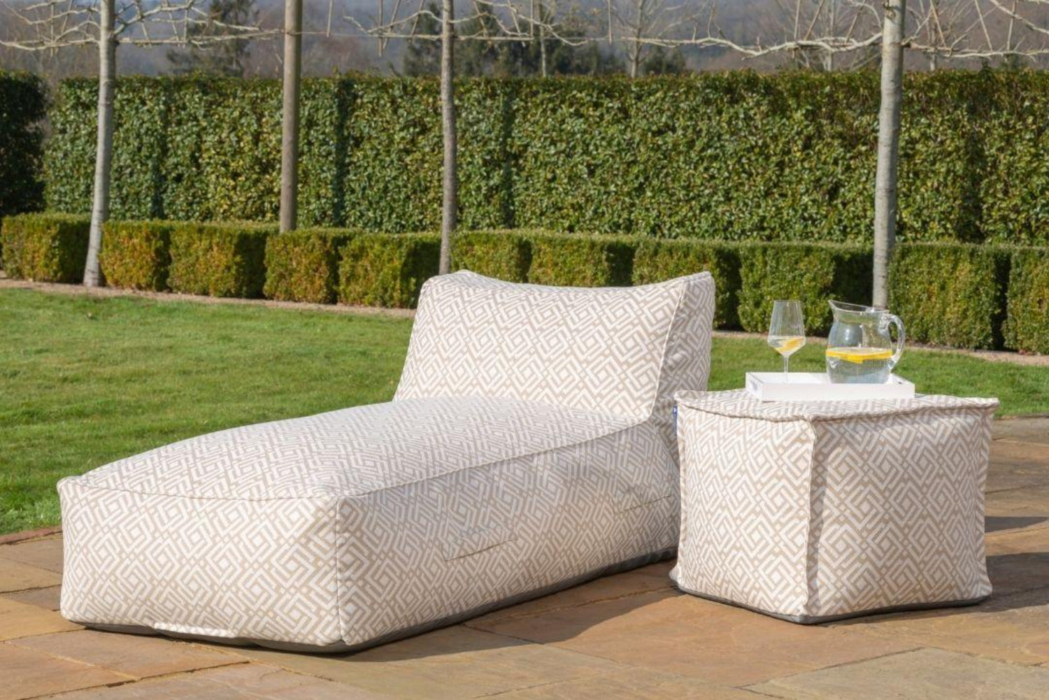 Outdoor bean bag sun lounger and matching side table in a garden setting, both upholstered in a light beige geometric fabric called sigma. The lounge is shown on a stone patio with a glass of wine and a pitcher on the side table, with a green hedge and lawn in the background.