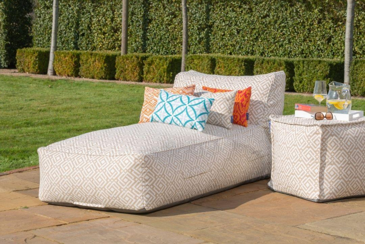 A luxury bean bag garden sun lounger with natural sigma patterned fabric, accompanied by a pouffe with a glass top, placed on a patio with greenery in the background. with Orange Tulip and Medina outdoor cushions.