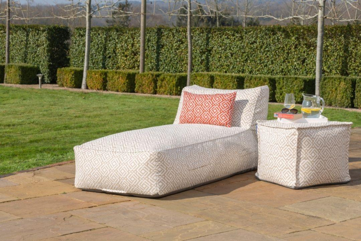 Outdoor bean bag sun lounger and matching pouffe in a light beige patterned fabric called sigma, styled with a matching paprika outdoor cushions in a garden setting.