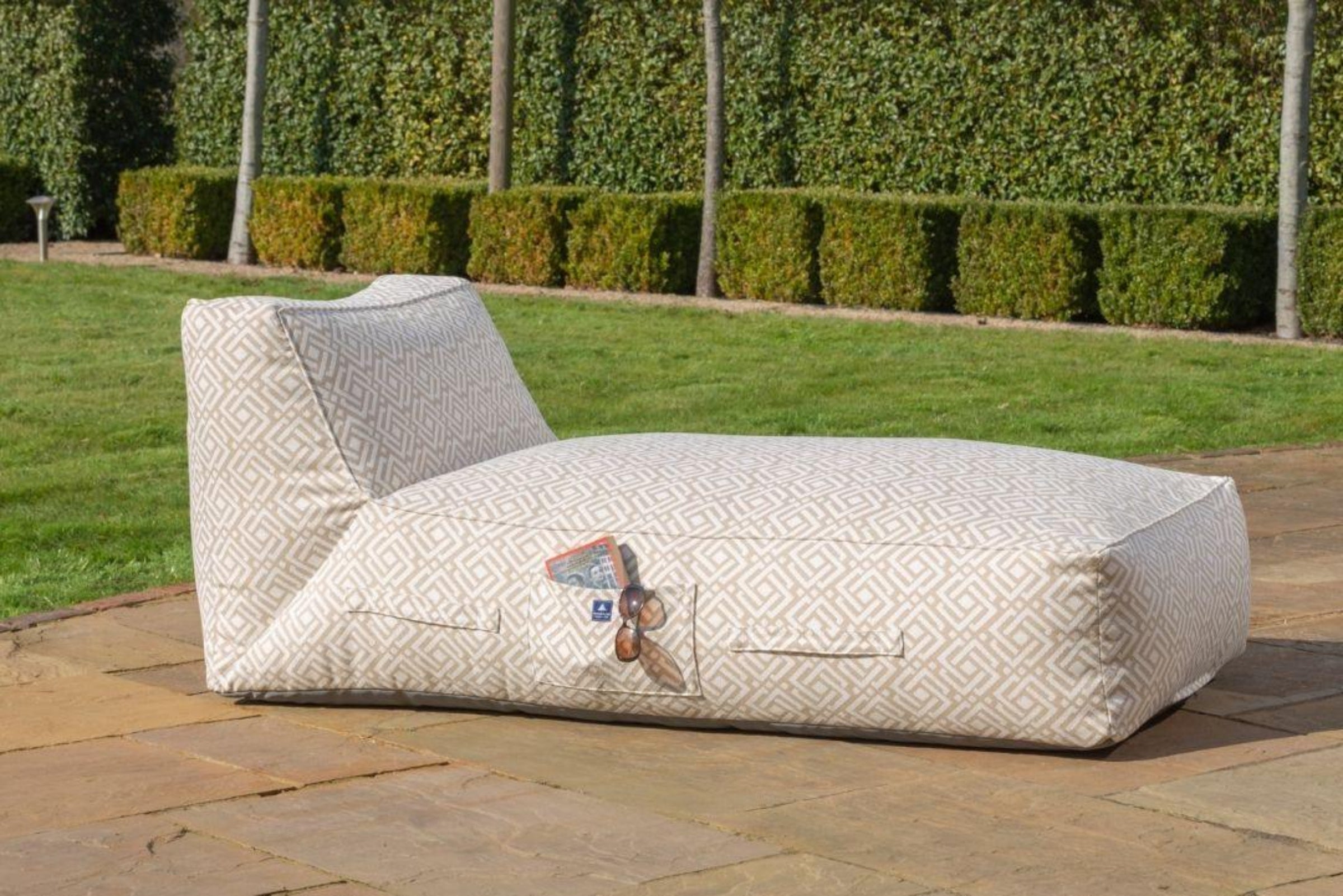 An outdoor bean bag sun lounger with a light beige and white geometric patterned fabric called sigma, placed on a stone patio in a garden setting.